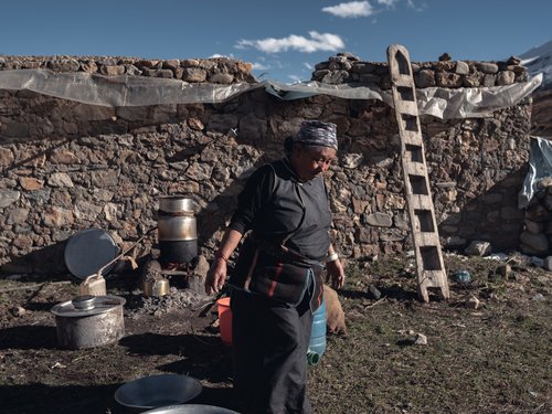 A woman tending an outdoor hearth by a dry-stone wall. At this altitude firewood is scarce; dung and juniper are the primary fuels.