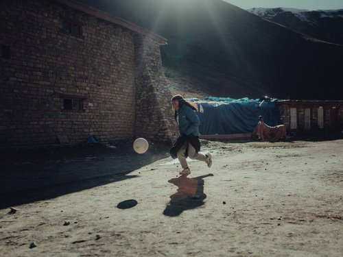 A child plays in bright October sun outside a stone building. At 3,900 m, solar irradiance on a clear day exceeds 1,000 W/m² — the resource the passive solar strategy is built on.